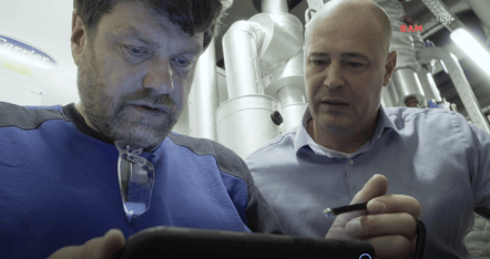 Twee mannen, één in een blauw shirt en de ander in een lichtblauw overhemd, kijken samen naar een smartphone in een industriële omgeving. Op de achtergrond zijn metalen leidingen zichtbaar.