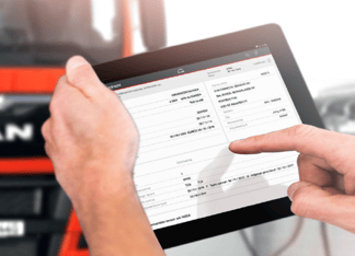 Een hand wijst op een tablet met een document zichtbaar, waarschijnlijk een vrachtbrief. Op de achtergrond is een deel van een vrachtwagen zichtbaar, met een feloranje kleur