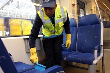 Een schoonmaker in fluorescerend veiligheidshesje en gele handschoenen veegt met een kleine handbezem tussen de blauwe stoelen in de coupé van een trein
