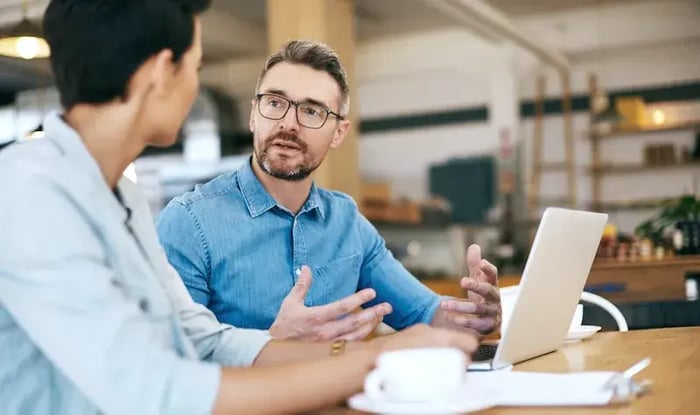 Een man legt iets uit terwijl hij tegenover een vrouw zit, met een laptop op tafel.