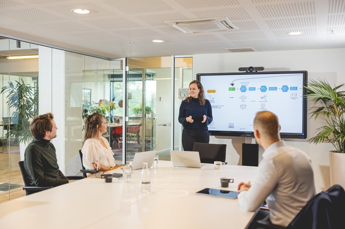 Een vrouw geeft een presentatie aan collega’s in een vergaderruimte met een groot scherm.