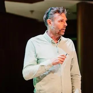 Portrait of Mark de Jong in a light colored shit with grey hair and a microfone on