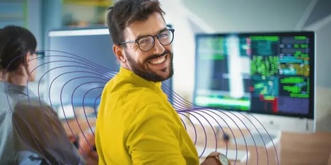 A white man with dark hair, a beard and glasses, in a yellow shirt, smiling in front of the camera behind his computer