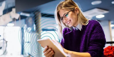 A white, blond hair lady, with glasses, in a purple cardigan, smiling at her tablet