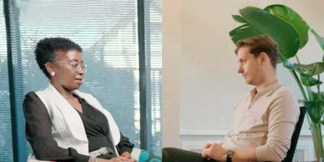 A woman with dark, short, frizzy hair and glasses talking to blond haired man