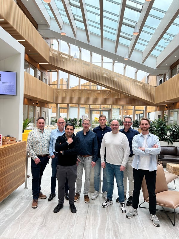 Groep van acht mensen die samen poseren in een grote, moderne lichte ruimte met glazen dak, houten balustrades en veel planten.