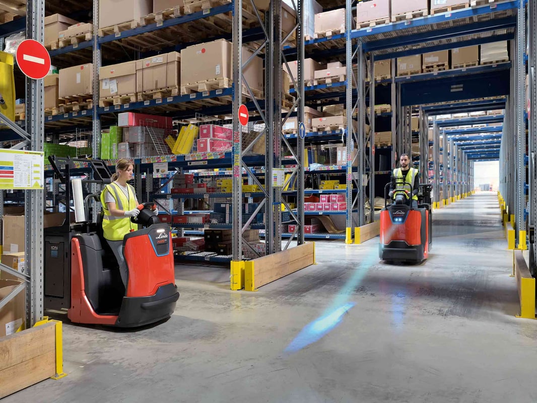 Twee medewerkers rijden met Linde-orderpickers door een magazijnpad met hoge stellingen vol dozen.