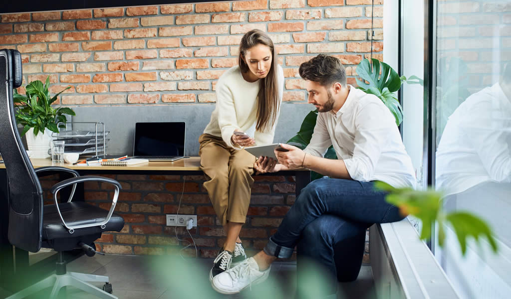Twee collega's bekijken samen een tablet in kantoor met bakstenen muur.
