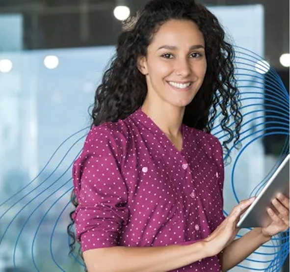 Eine weiße Dame mit braunen, lockigen langen Haaren, die vor der Kamera lächelt, in einer magentafarbenen Bluse mit weißen Punkten und einem Tablet in der Hand.