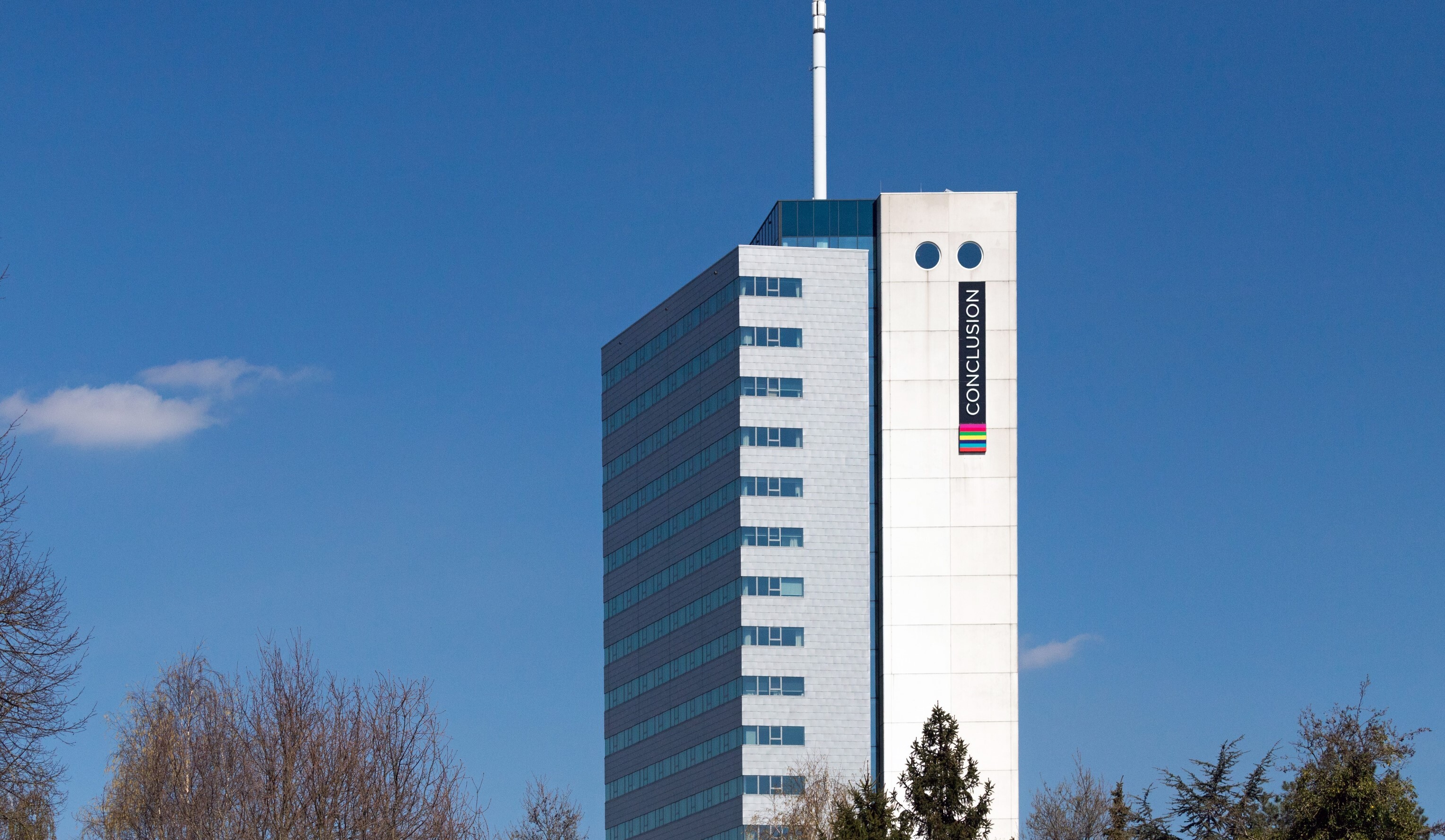 Hoog modern kantoorgebouw met het logo van Conclusion op de gevel, omgeven door bomen en gezet tegen een helderblauwe lucht.