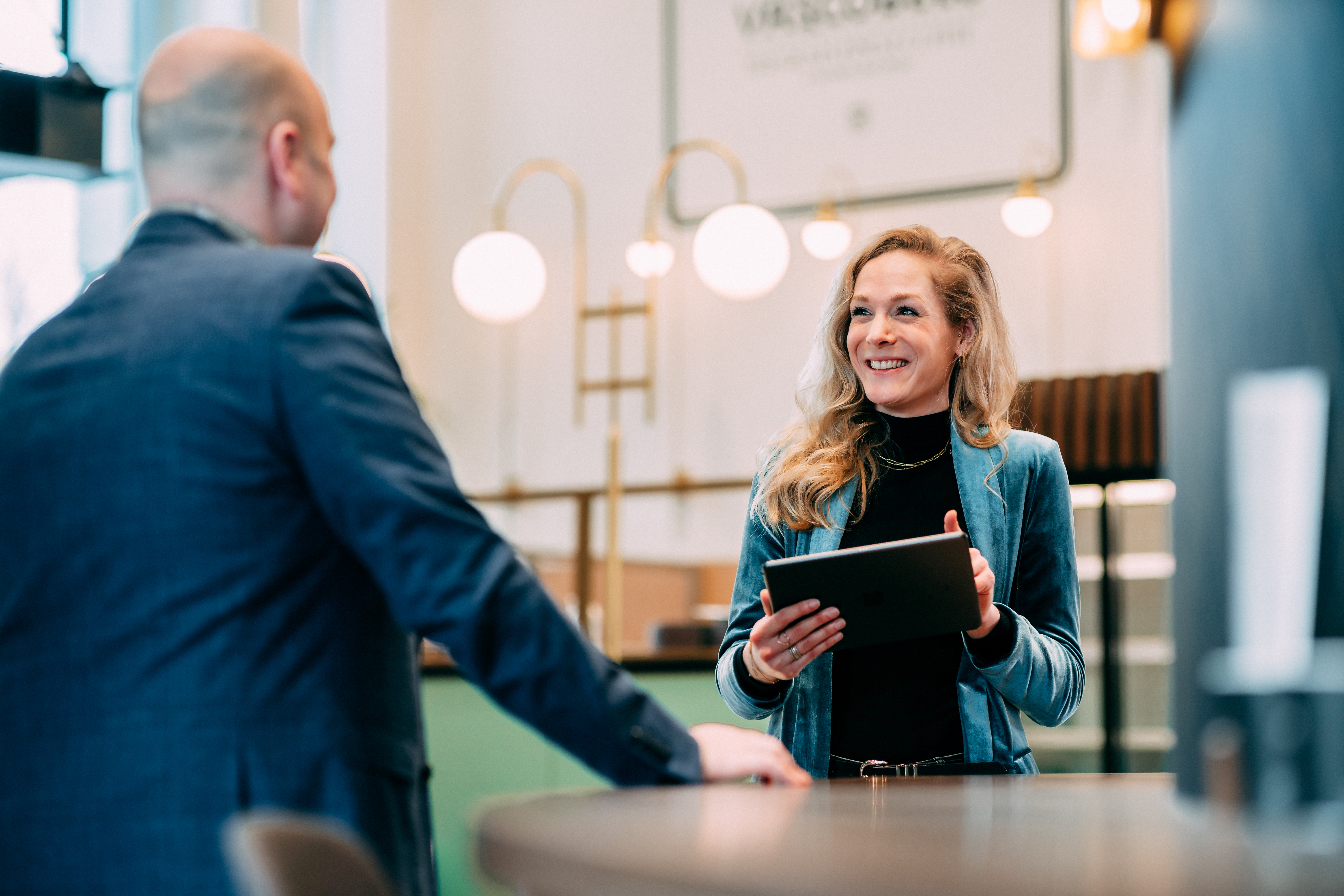 Vrouw die glimlachend met een tablet in haar handen een gesprek voert met een man in een modern ingerichte ruimte met warme verlichting en een professionele sfeer.
