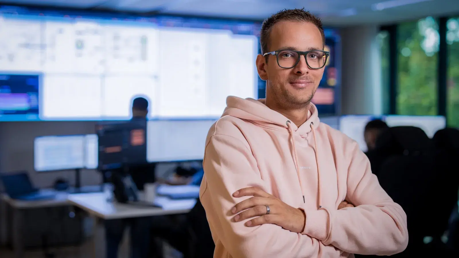 Man met armen over elkaar met monitors op de achtergrond