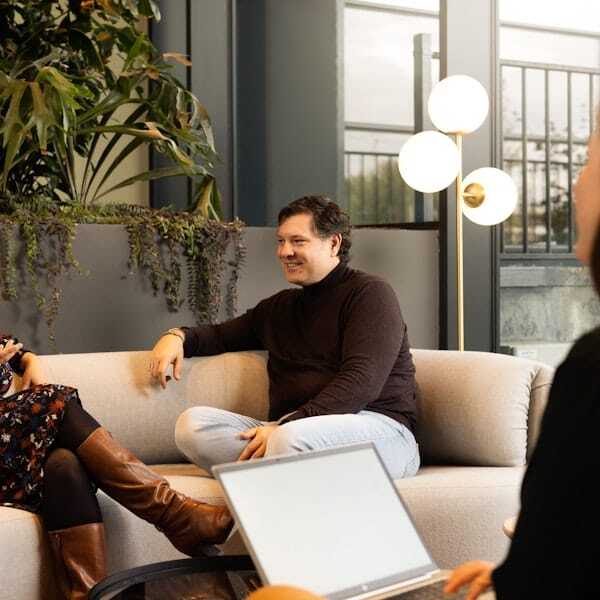 Mannelijk collega zit op een bank en kijkt vriendelijk.