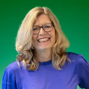 Portret van een lachende vrouw met blond haar en bril voor een groene achtergrond.