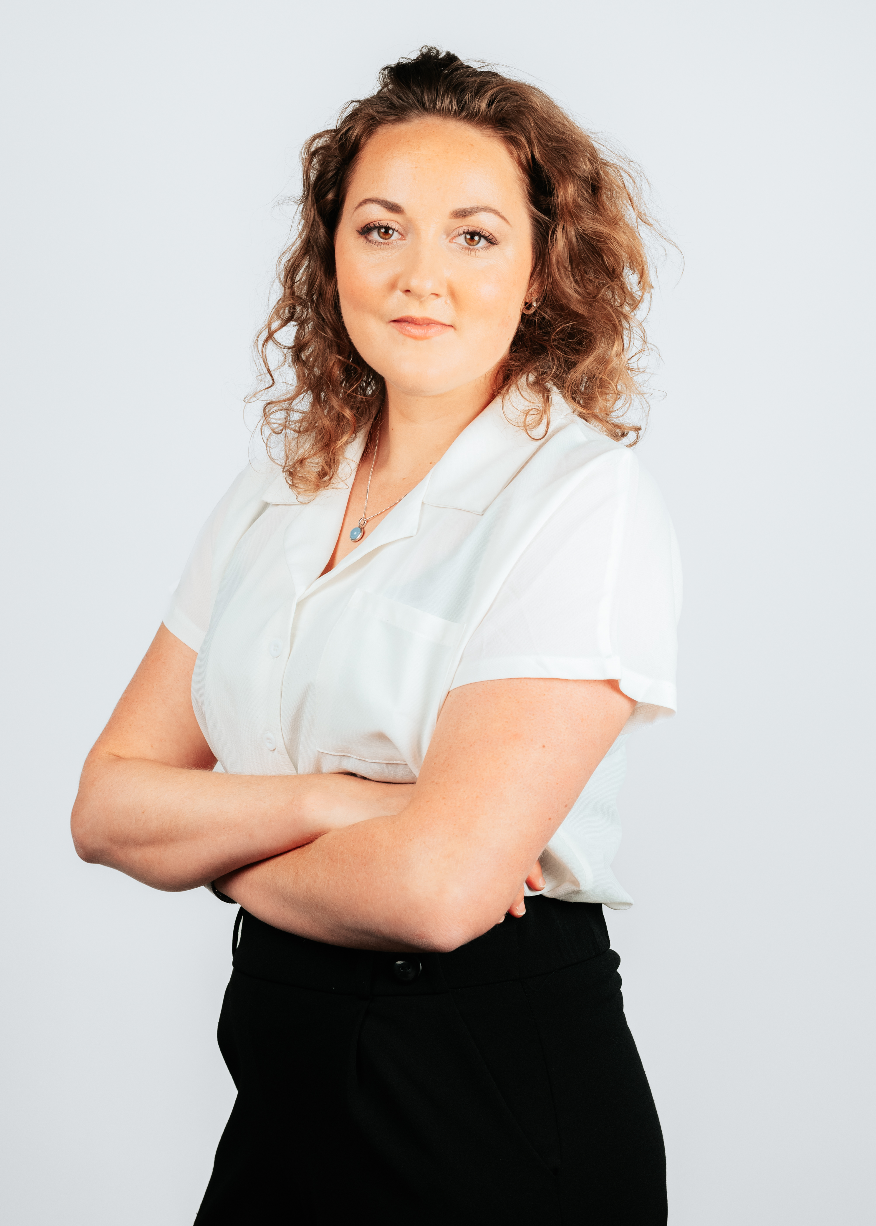 Vrouw met krullend haar en een zelfverzekerde houding, gekleed in een witte blouse en zwarte broek, poserend tegen een lichte achtergrond.