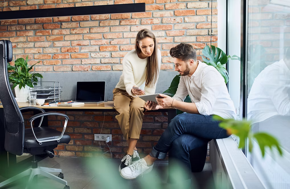 Twee collega's overleggen samen over een tablet in een modern kantoor met bakstenen muur.